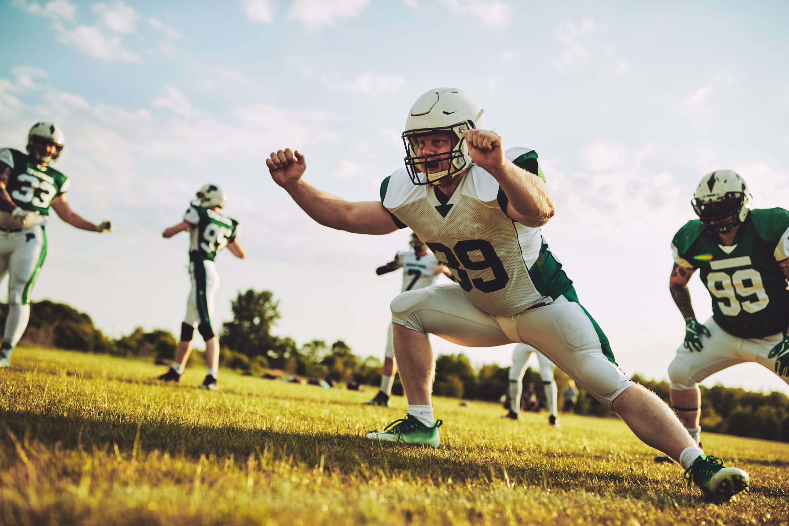 American football players in helmets and pads practicing defensive drills on a sunny field, with a focus on a player in a stance ready for the play.