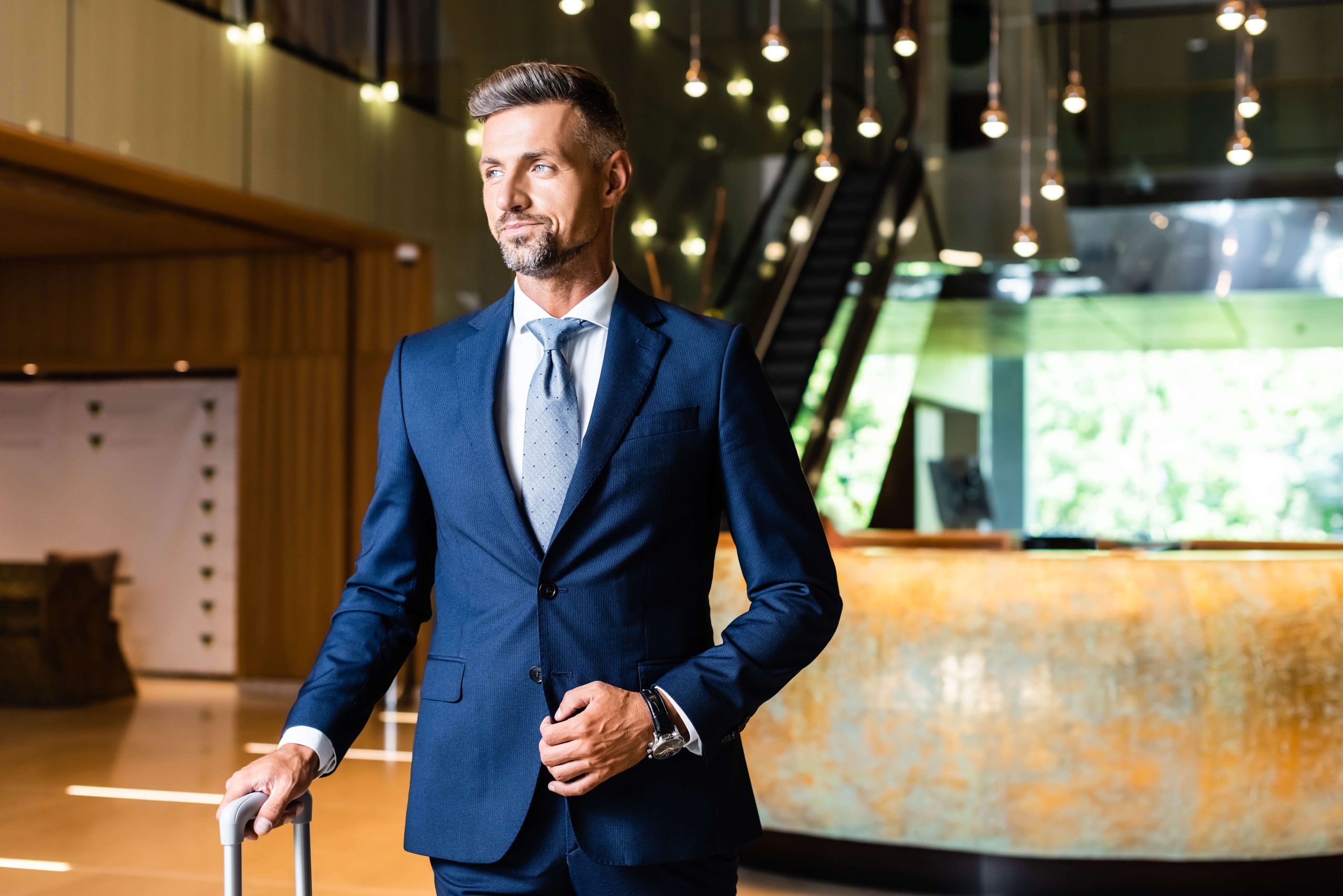 A confident middle-aged businessman in a pressed and tailored blue suit with a light blue tie and a wristwatch, holding a suitcase handle, looking away thoughtfully in a modern hotel lobby with hanging lights and a staircase in the background.