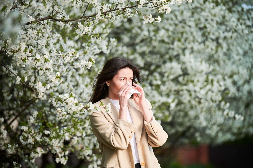 A woman standing outside in a blooming garden, blowing her nose with a tissue, likely suffering from seasonal allergies. She is wearing a light beige blazer and a white shirt, with a backdrop of densely flowering trees indicating springtime.