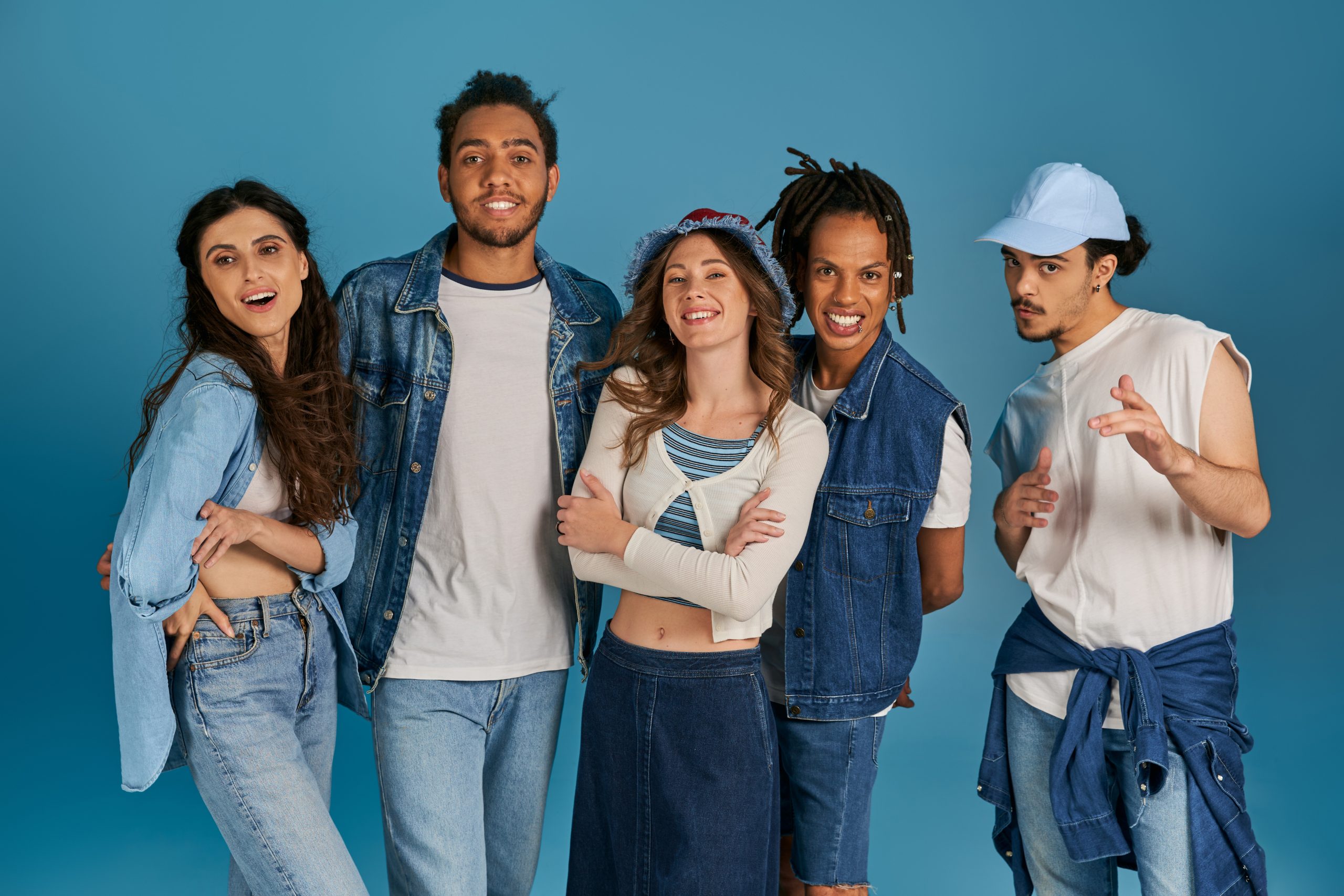 A group of multicultural friends smiling and posing together, wearing trendy denim outfits and casual wear against a blue background.