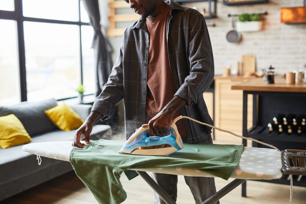 A man attentively ironing a green shirt on an ironing board in a cozy, well-lit home setting, with steam rising from the iron.