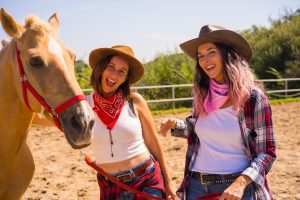 Two happy women in cowgirl attire with cowboy hats and bandanas smiling beside a palomino horse with a red halter.
