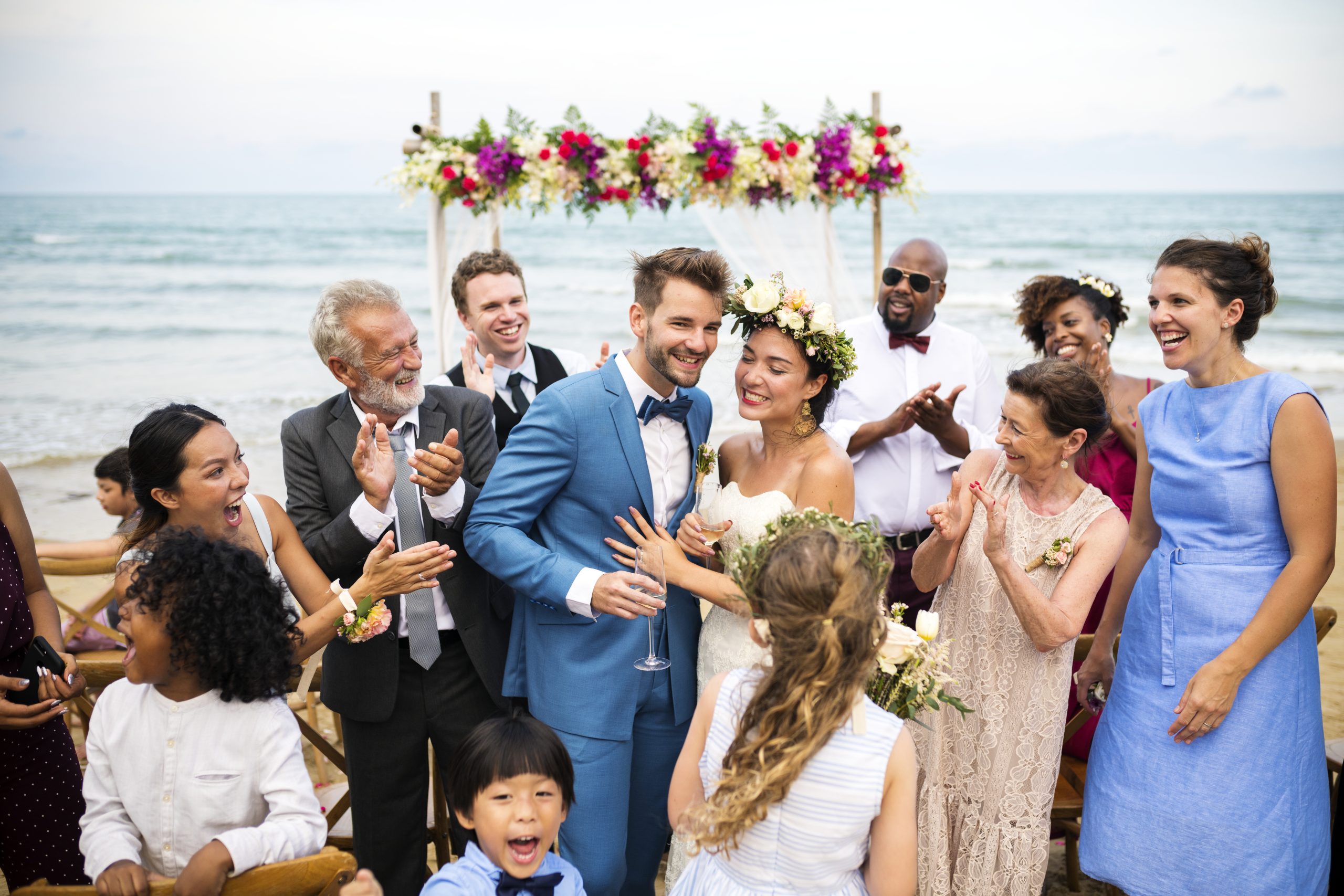 A joyful wedding scene on the beach with a diverse group of guests clapping and celebrating a young couple in their wedding attire.