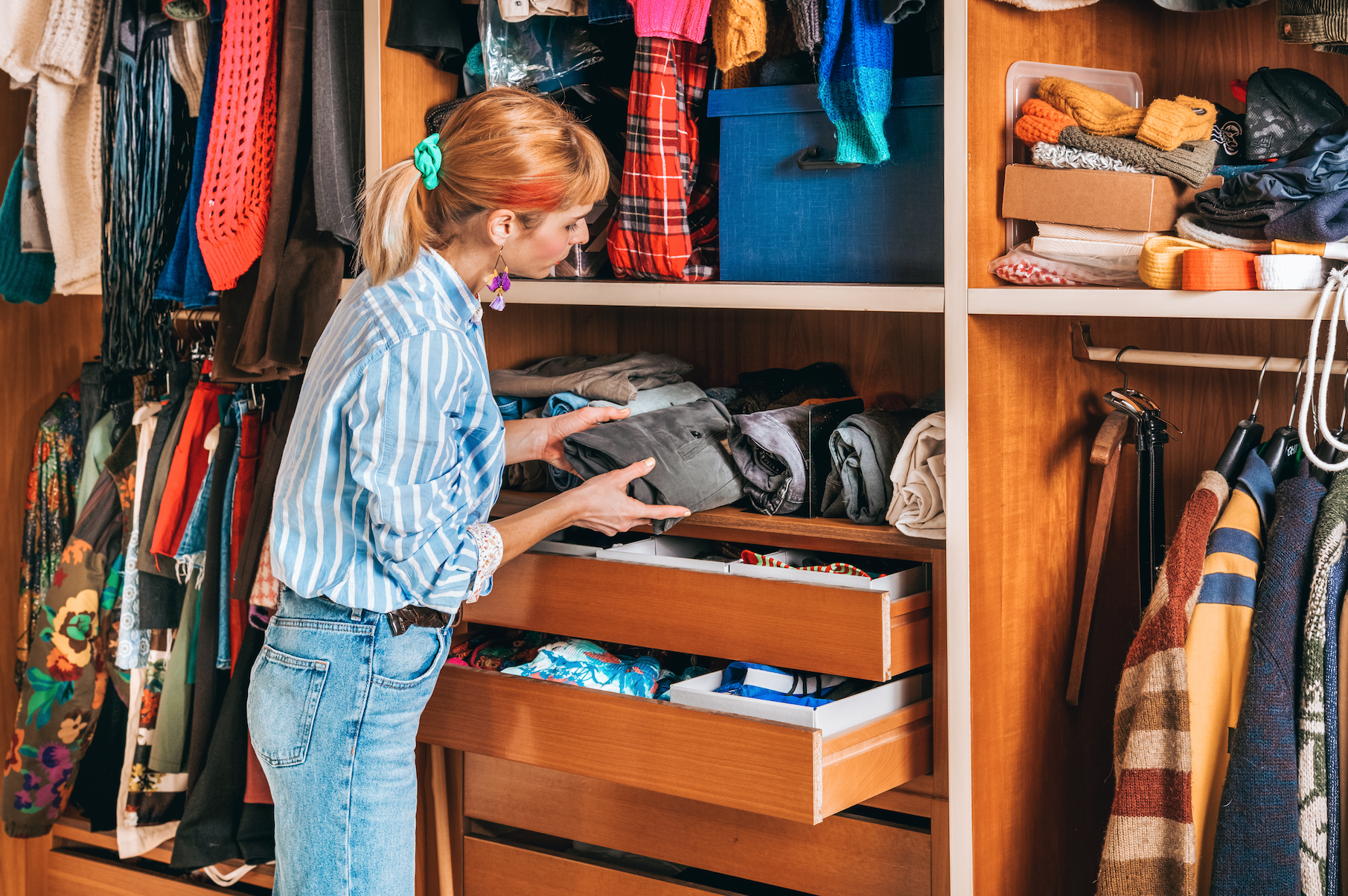 Woman organizing clothes in a well-stocked wooden wardrobe, examining various garments and accessories.