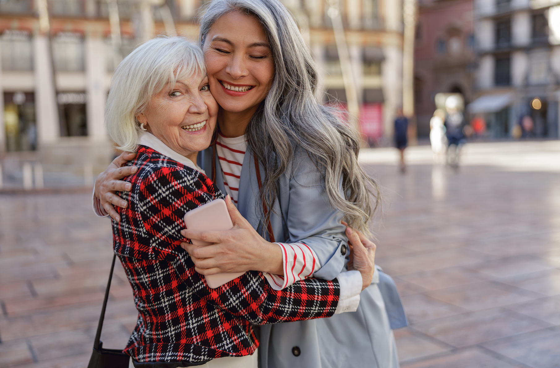 Happy embrace between an elderly woman and her adult daughter, smiling and holding a smartphone, in a lively urban square.