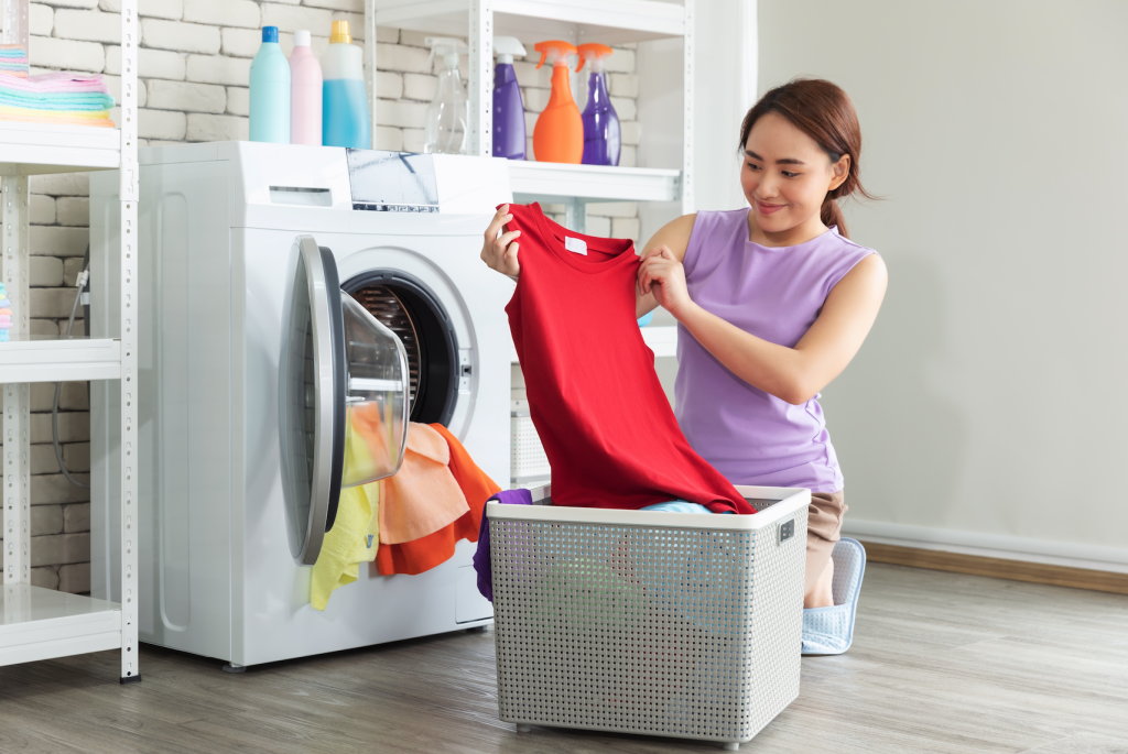 Woman smiling as she examines a red sleeveless top while doing laundry in a modern laundry room with shelves of supplies and a washing machine.