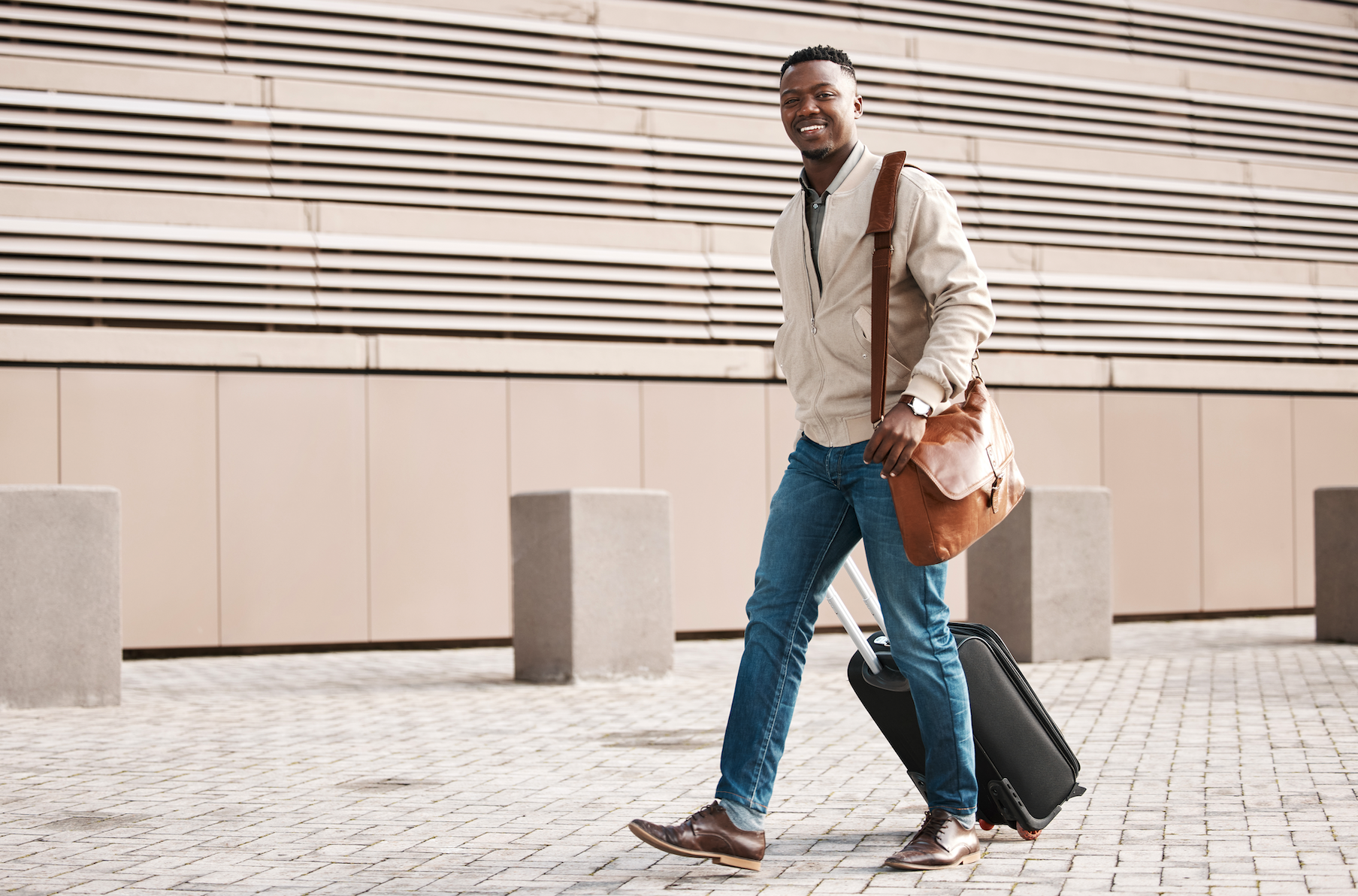 Smiling man walking with a suitcase and a leather shoulder bag, dressed in a casual sweater and jeans, in front of a modern building.