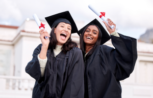 Two female graduates in black caps and gowns smiling and holding diplomas.