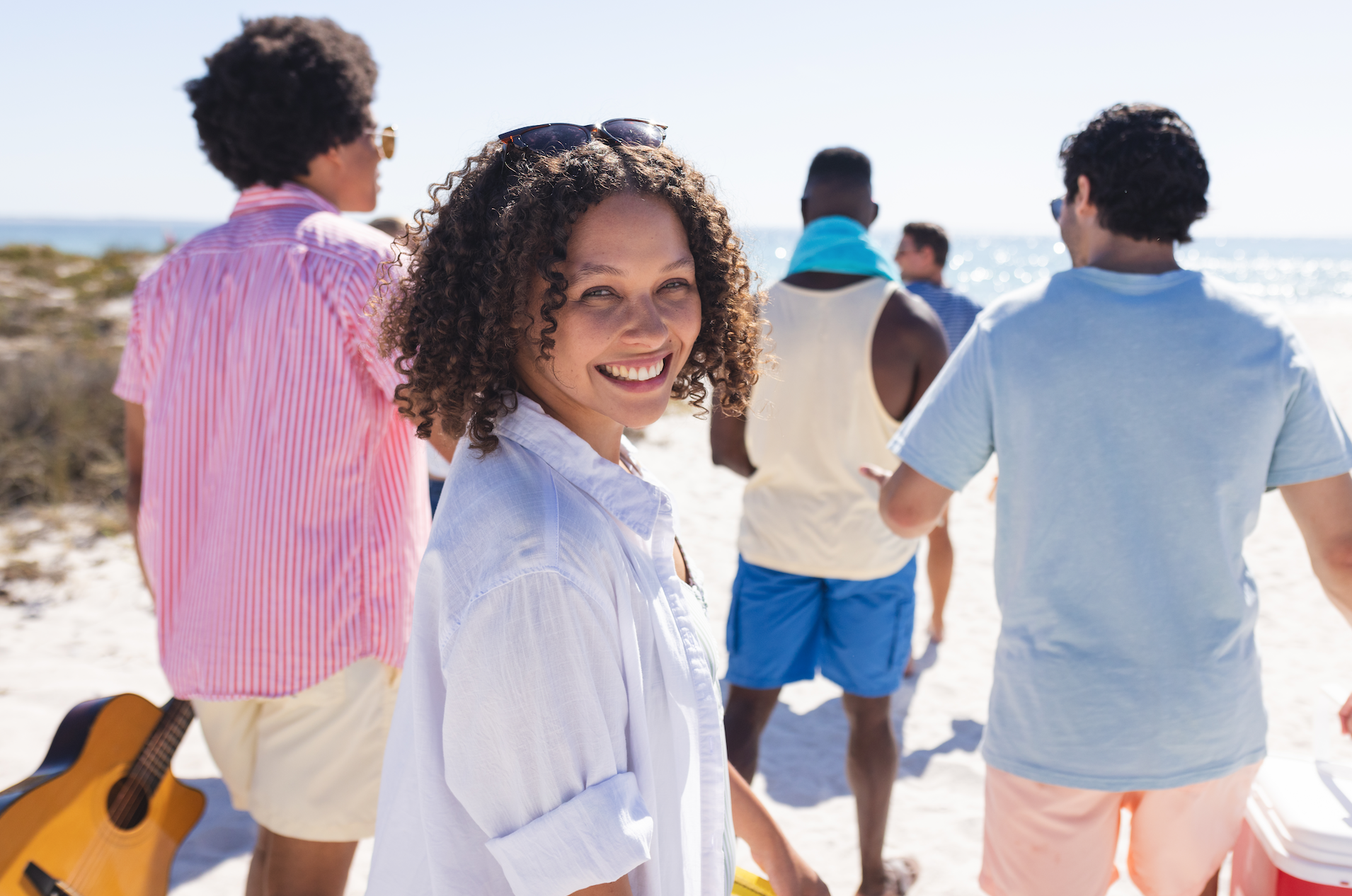 A group of friends enjoying a sunny day at the beach, with a smiling woman in the foreground and a guitar in the background.