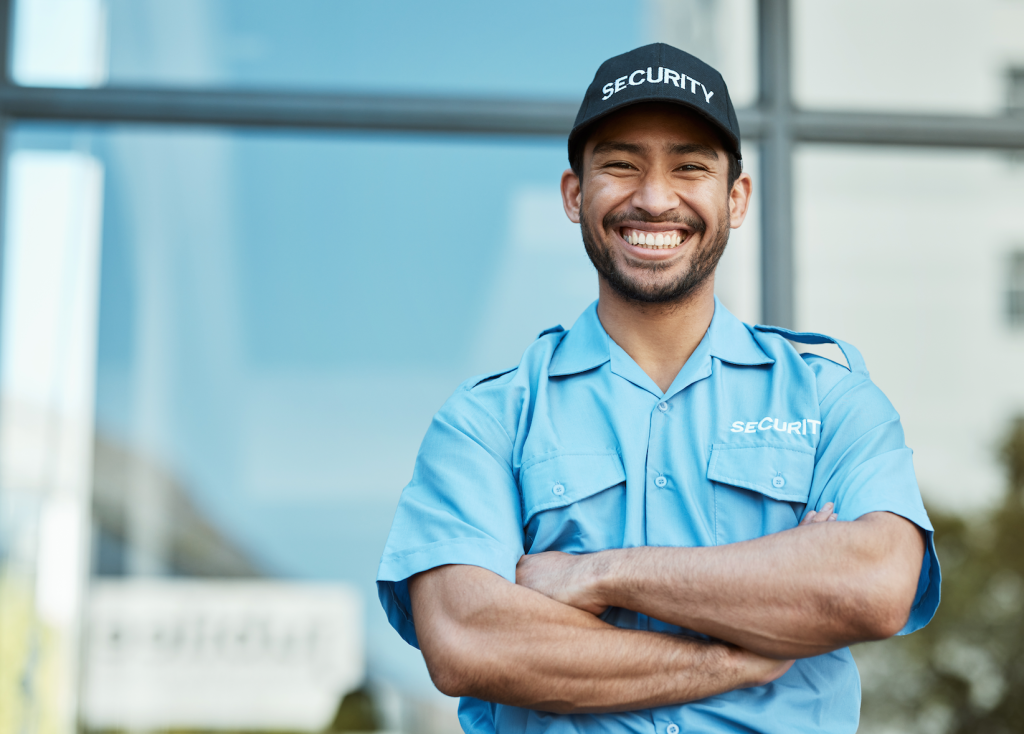 A smiling security guard in a blue uniform and black cap with arms crossed, standing in front of a modern building.