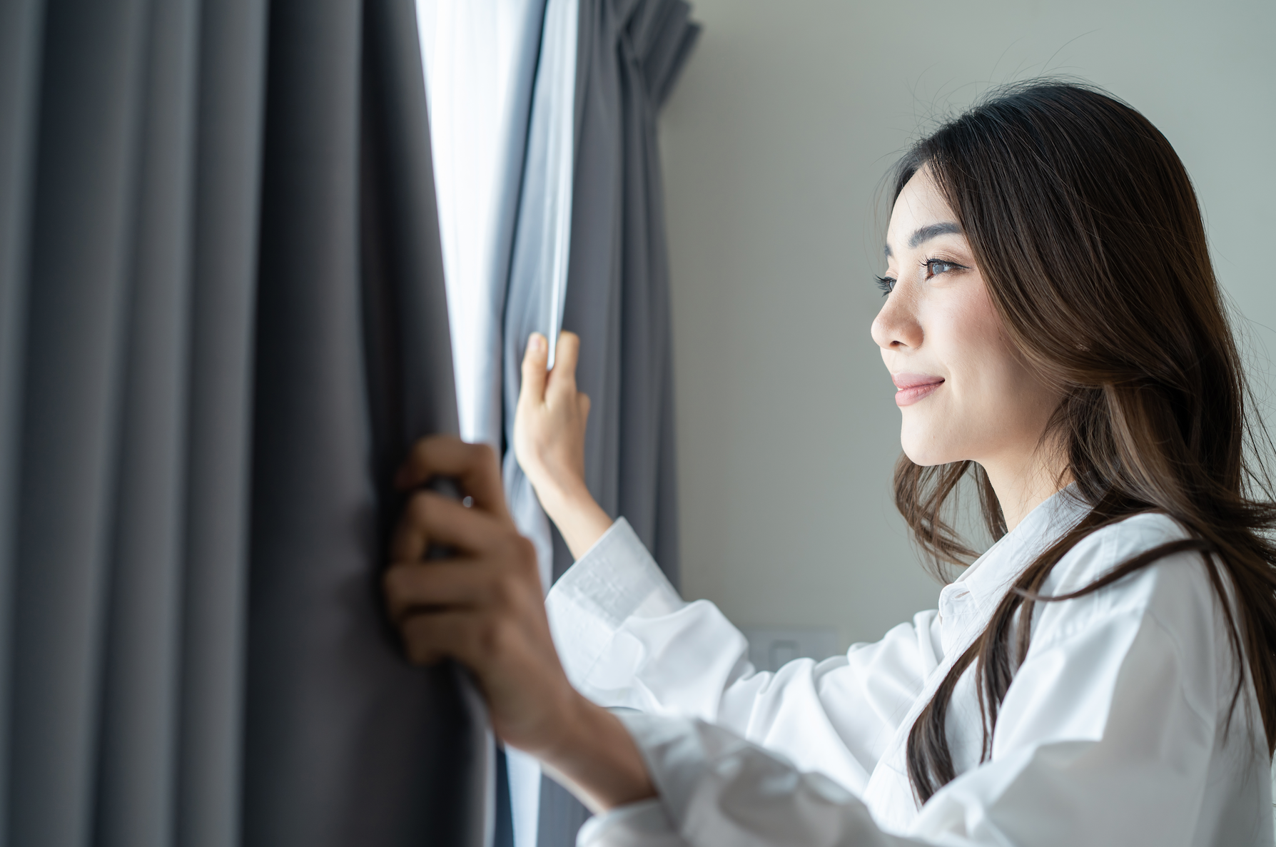 A woman smiling as she opens gray curtains, letting natural light into the room.